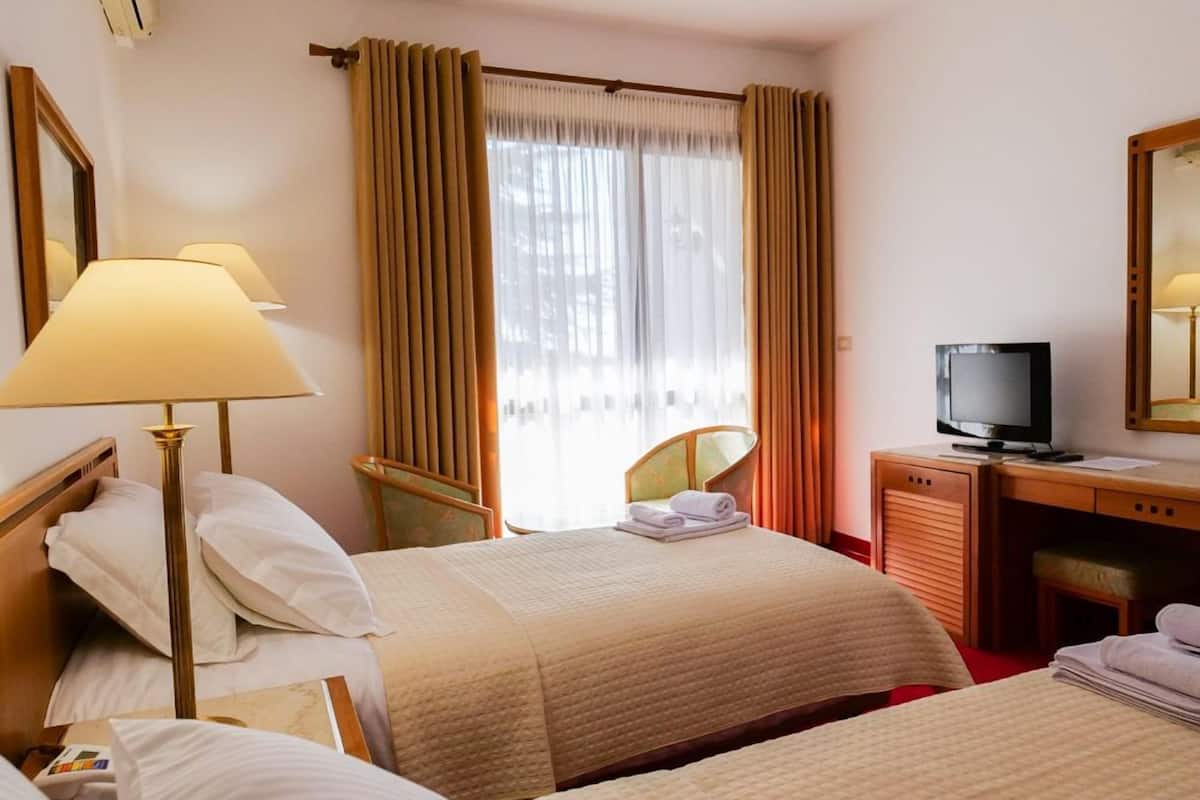 Ein Hotelzimmer mit zwei Betten, einem Schreibtisch mit Fernseher, einem Sessel und einem Fenster mit Vorhängen.
