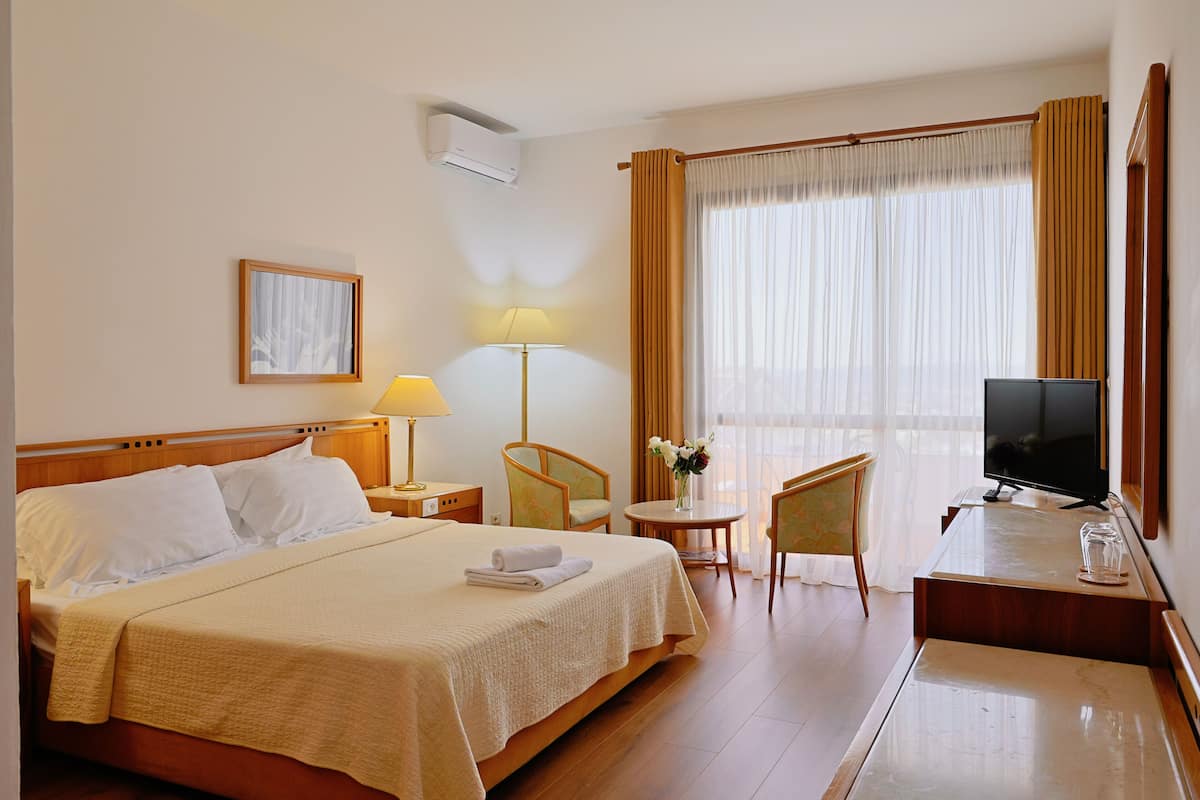 Ein Hotelzimmer mit Bett, Fernseher, Schreibtisch mit Stuhl, Lampe, Fenster mit Vorhängen und einem Bild in einem Rahmen an der Wand.