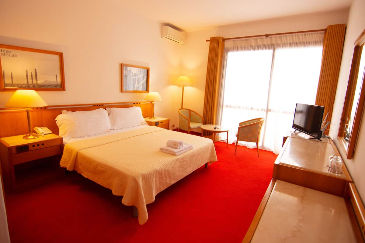 Ein Hotelzimmer mit einem großen Bett, Nachttischlampen, einem Schreibtisch mit Fernseher und einem Vorhangfenster.