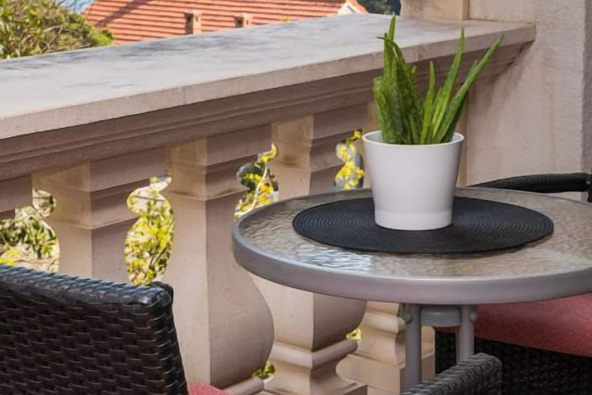 Un balcón con una mesa redonda, dos sillas de mimbre, una planta en maceta y vistas al mar y edificios.