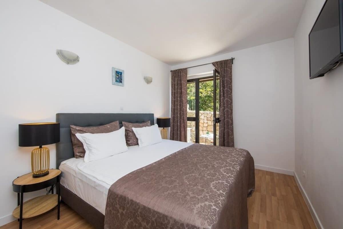 Un dormitorio con cama, mesita de noche, lámpara, televisor en la pared y ventana con cortinas.