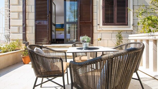 An outdoor seating area with wicker chairs and a table, a potted plant, and a building entrance with a glass door and shutters. - Villa Dard (Dubrovnik)