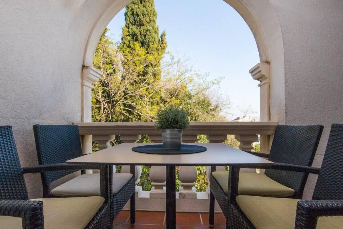 Un patio con una mesa, cuatro sillas, un arco y vistas a árboles y un edificio.