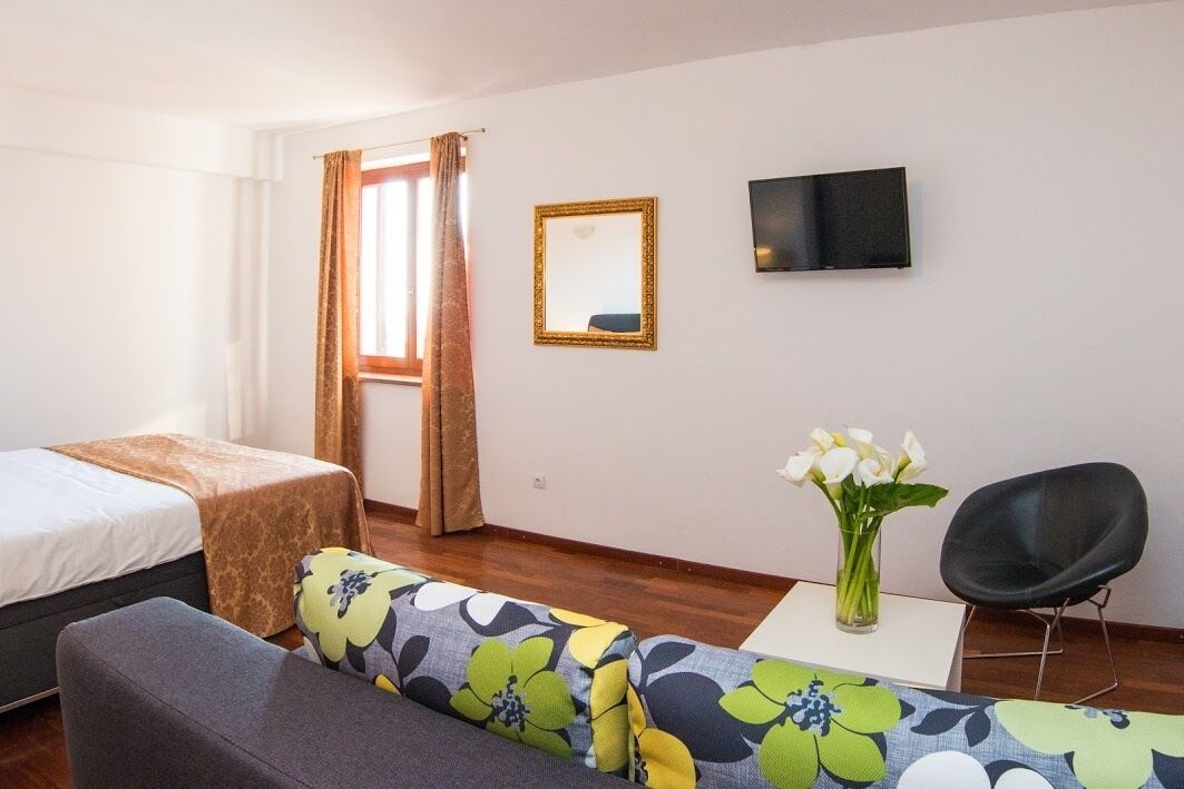 A hotel room with a bed, a sofa, a small table with a vase of flowers, a chair, a TV, and a window with curtains.