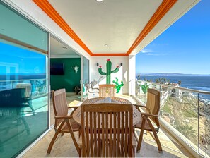 A balcony with a wooden table and chairs, a cactus mural, and a view of the ocean. - JustViews-LasOlasCondo-Pools&Jacuzzis-2BR-Rosarito (Playas Rosarito)