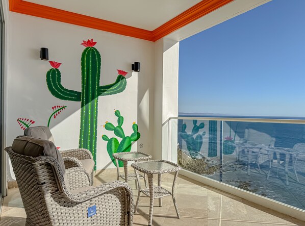 A balcony with wicker furniture, a large cactus mural, and a view of the ocean. - JustViews-LasOlasCondo-Pools&Jacuzzis-2BR-Rosarito (Playas Rosarito)