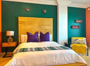 A bedroom with a large bed, a wooden headboard, a nightstand with a lamp, and a chair with a yellow blanket. - JustViews-LasOlasCondo-Pools&Jacuzzis-2BR-Rosarito (Playas Rosarito)