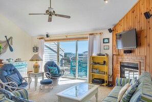 A living room with a ceiling fan, a TV mounted on the wall, a fireplace, and a balcony with a view of buildings. - La Mar: Semi-oceanfront Townhome, Shared Pool (Nags Head)