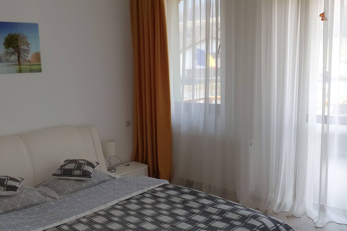 A bedroom with a bed, pillows, a bedside table with a lamp, and a window with sheer curtains.