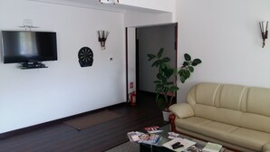 A living room with a flat-screen TV, a dartboard, a fire extinguisher, a potted plant, and a beige sofa. - Pensiunea Caraiman (Codlea)