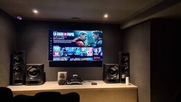 Sistema de cinema em casa com uma TV de tela plana grande fixada na parede, dois alto-falantes grandes de cada lado e um console de jogos à frente.