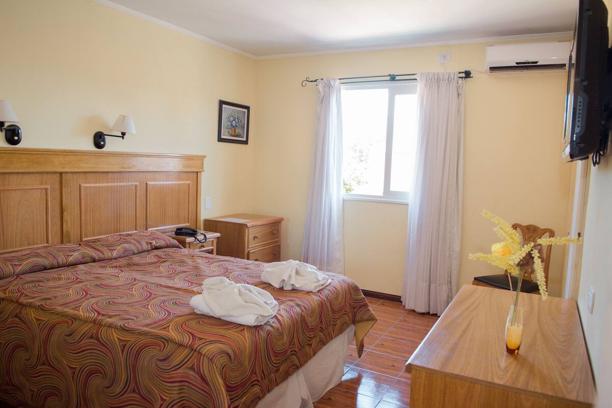 Une chambre d’hôtel comprenant un lit, une table de chevet, une commode, une fenêtre avec des rideaux et un téléviseur à écran plat fixé au mur.