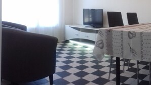 Una habitación con suelo a cuadros, una mesa de comedor con mantel floral, un televisor sobre un mueble blanco y un cuadro en la pared.