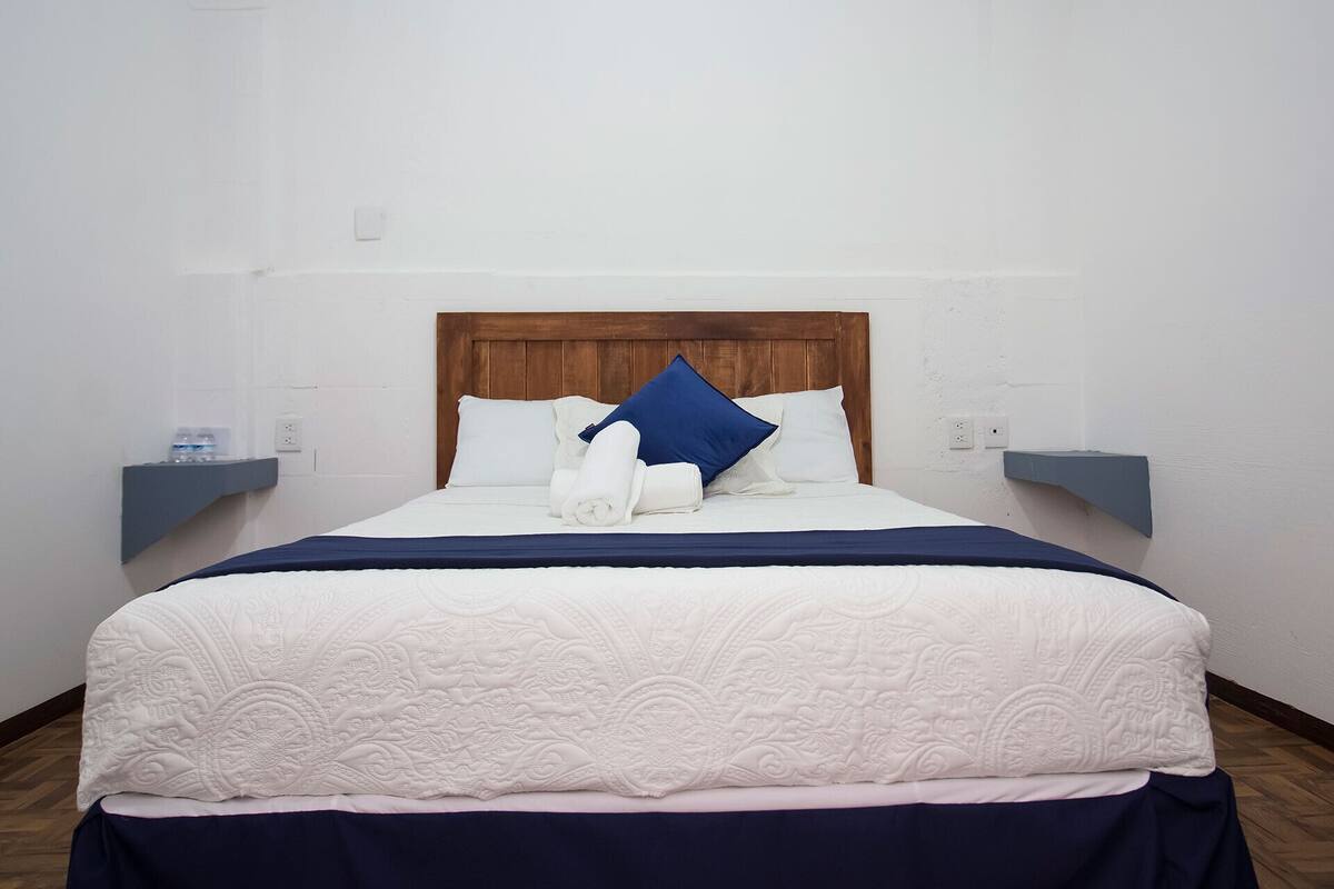 A neatly made bed with a wooden headboard, white linens, and a blue pillow. There are two bedside tables with water bottles on them.