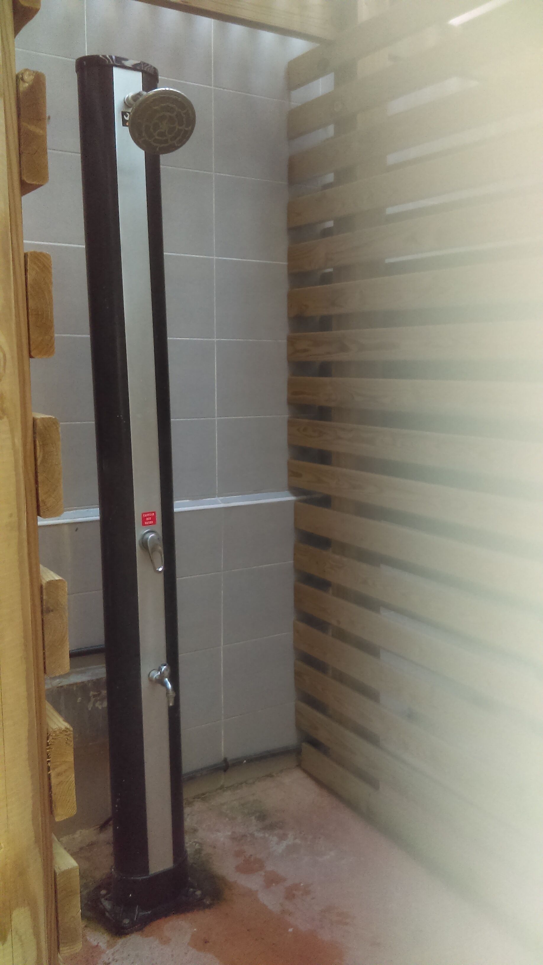 A shower stall with a vertical drain, a metal handle, and a red sticker on the wall.