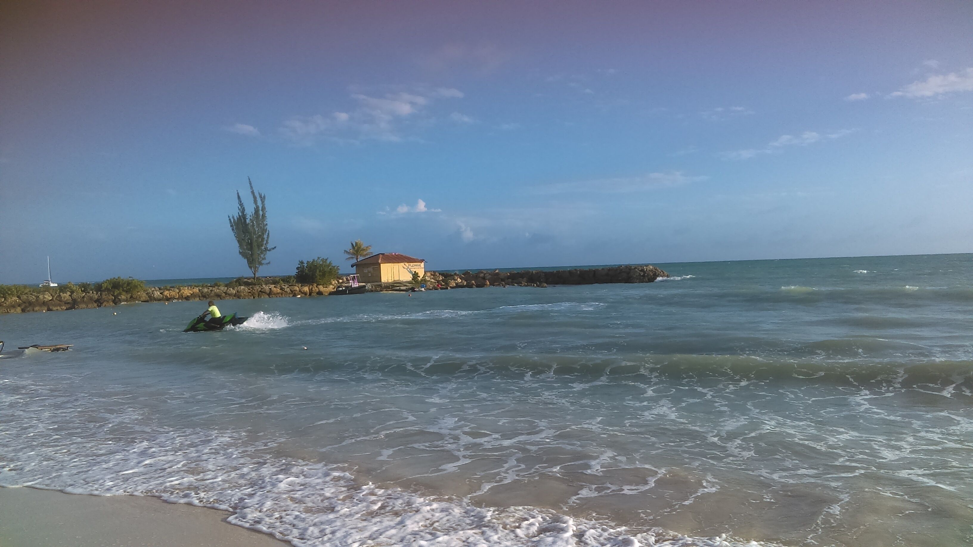 Una platja amb un pontó rocos i un edifici amb teulada vermella, amb una persona conduint un esquí aquàtic.