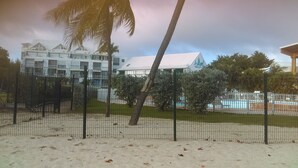 A hotel with a pool, surrounded by a fenced area with palm trees and a sandy ground. - Charming duplex with no water shut-off (Le Gosier)
