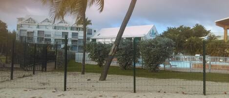 A hotel with a pool, surrounded by a fenced area with palm trees and a sandy ground.