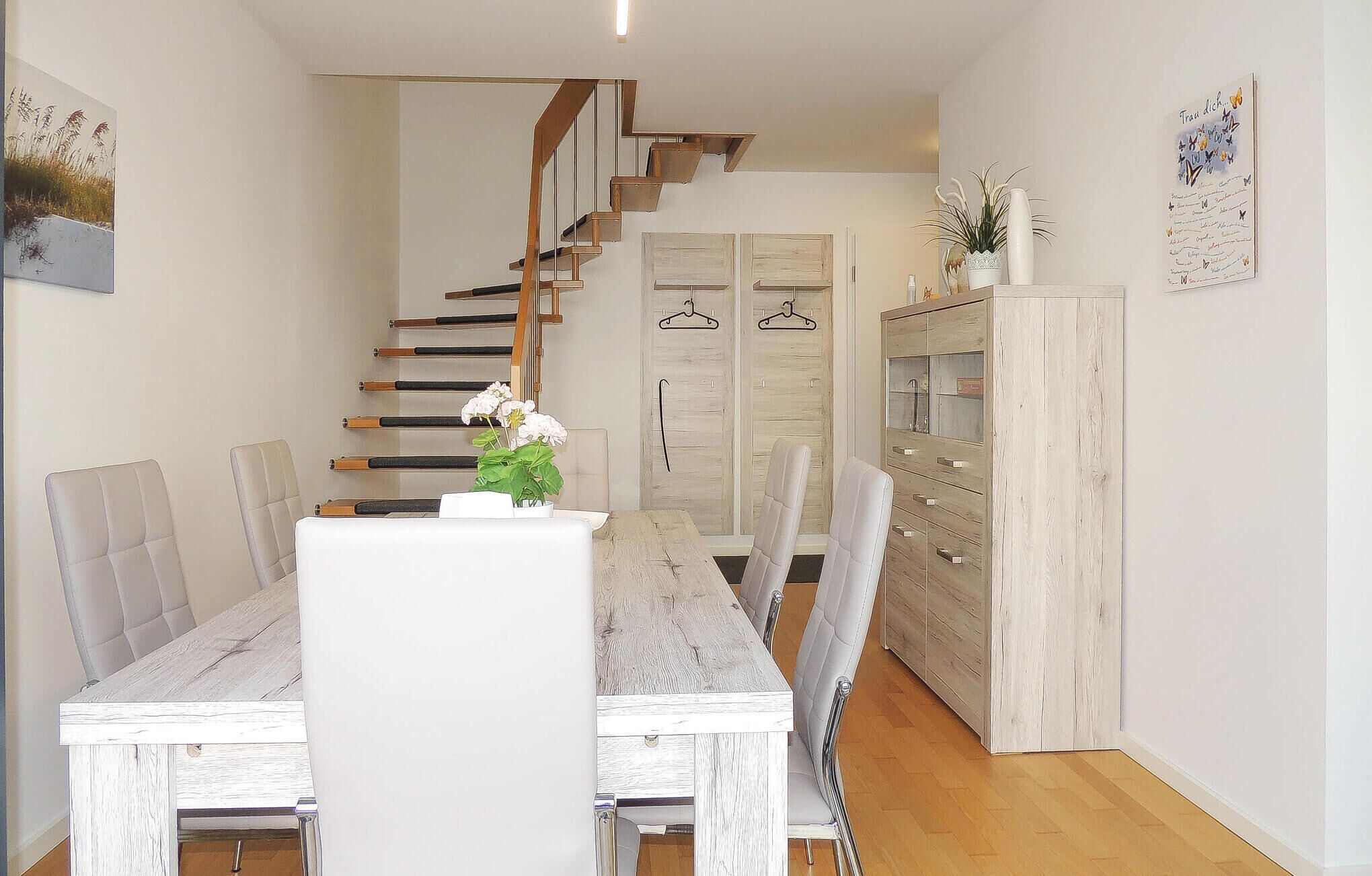 Un interior moderno con una escalera de madera, una zona de comedor con sillas blancas y una mesa de madera, y un armario con puertas de cristal.