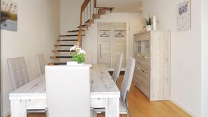 Un interior moderno con una escalera de madera, una zona de comedor con sillas blancas y una mesa de madera, y un armario con puertas de cristal.