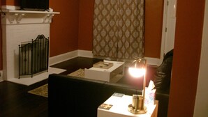 A living room with a fireplace, a television on a shelf, a patterned curtain, and a table with a lamp and a box.