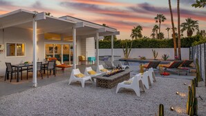 An outdoor patio with a fire pit, white lounge chairs, and a dining area with a table and chairs. - For The Record (Palm Springs)