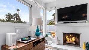 A modern living room with a fireplace, a flat-screen TV, and a view of palm trees. - For The Record (Palm Springs)