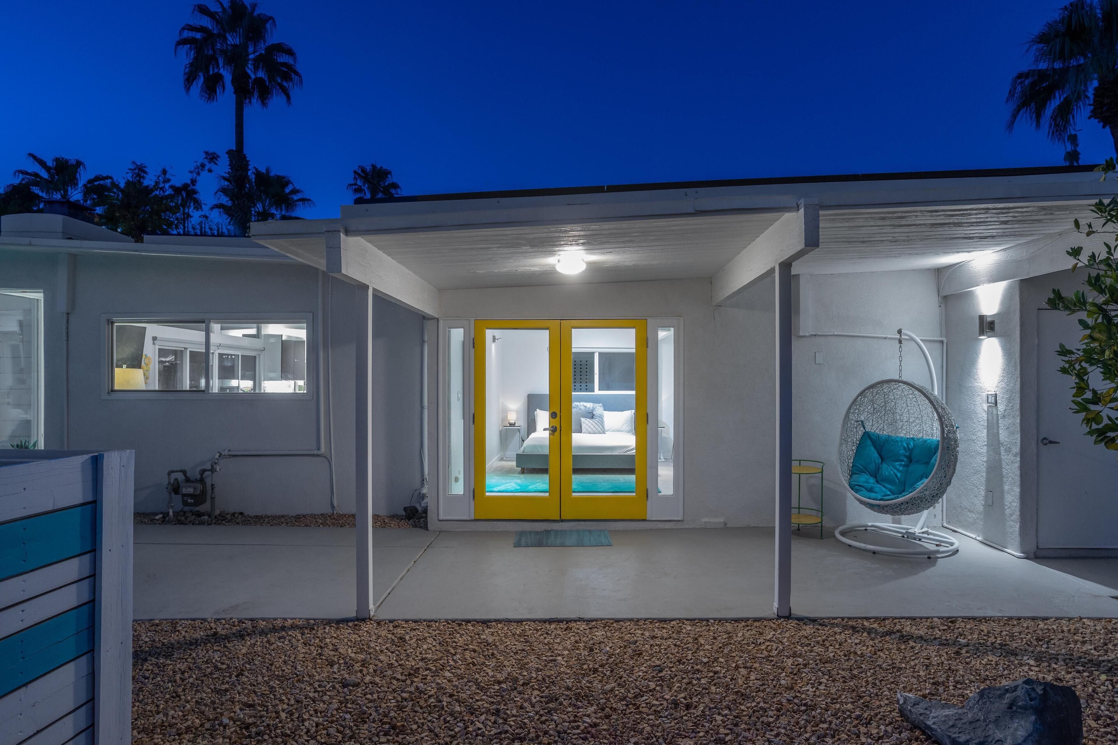 A modern house with a yellow door, a hanging chair, and a visible bedroom with a bed and nightstand.