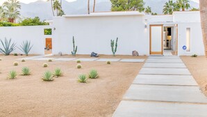 A modern house with a concrete pathway leading to the entrance, surrounded by cacti and palm trees, with mountains in the background. - For The Record (Palm Springs)
