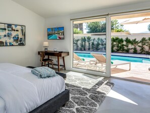 A bedroom with a bed, a desk, a chair, a TV, and a view of a swimming pool. - Movie Colony Palms (Palm Springs)