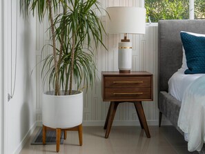 Un dormitorio con una cama, una mesita de noche con lámpara, una planta en maceta y otra mesita auxiliar.