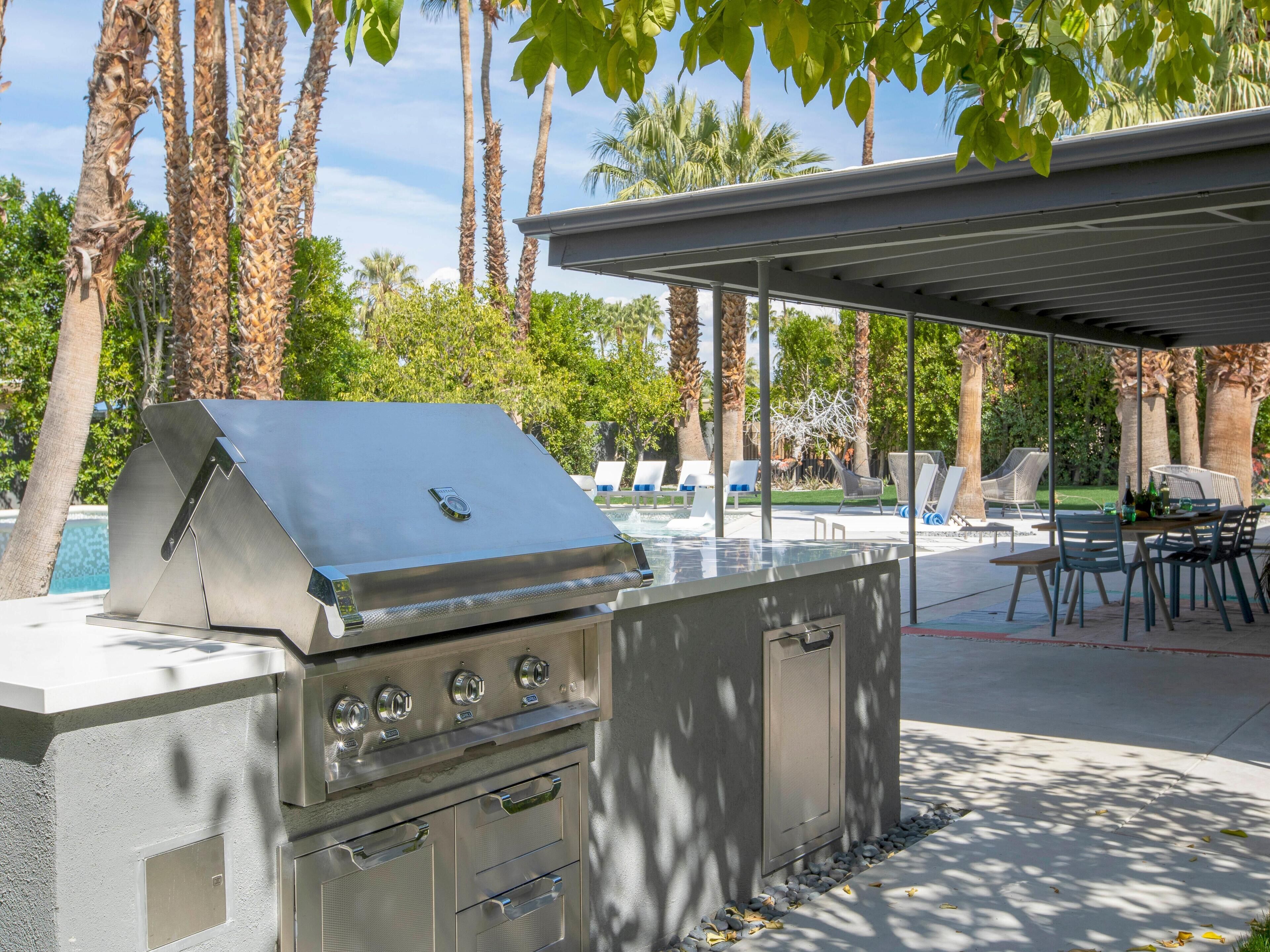 An outdoor kitchen with a grill, built-in drawers, and a covered dining area with chairs and a table.