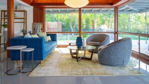 A modern living room with a blue sofa, round coffee table, and a stone accent chair. A wooden ceiling and large windows provide natural light and views of the outdoors.