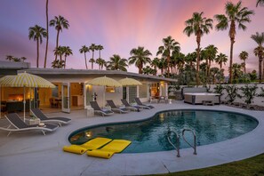 A pool area with lounge chairs and umbrellas, surrounded by palm trees and a building with a modern design.
