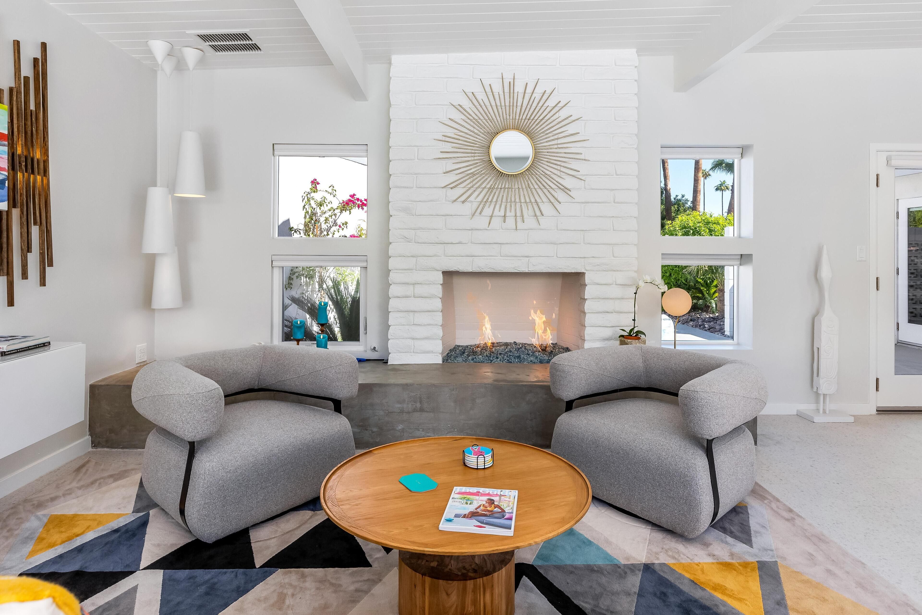 Ein modernes Wohnzimmer mit Kamin, zwei grauen Sesseln, einem runden Couchtisch und einem Teppich mit geometrischem Muster.