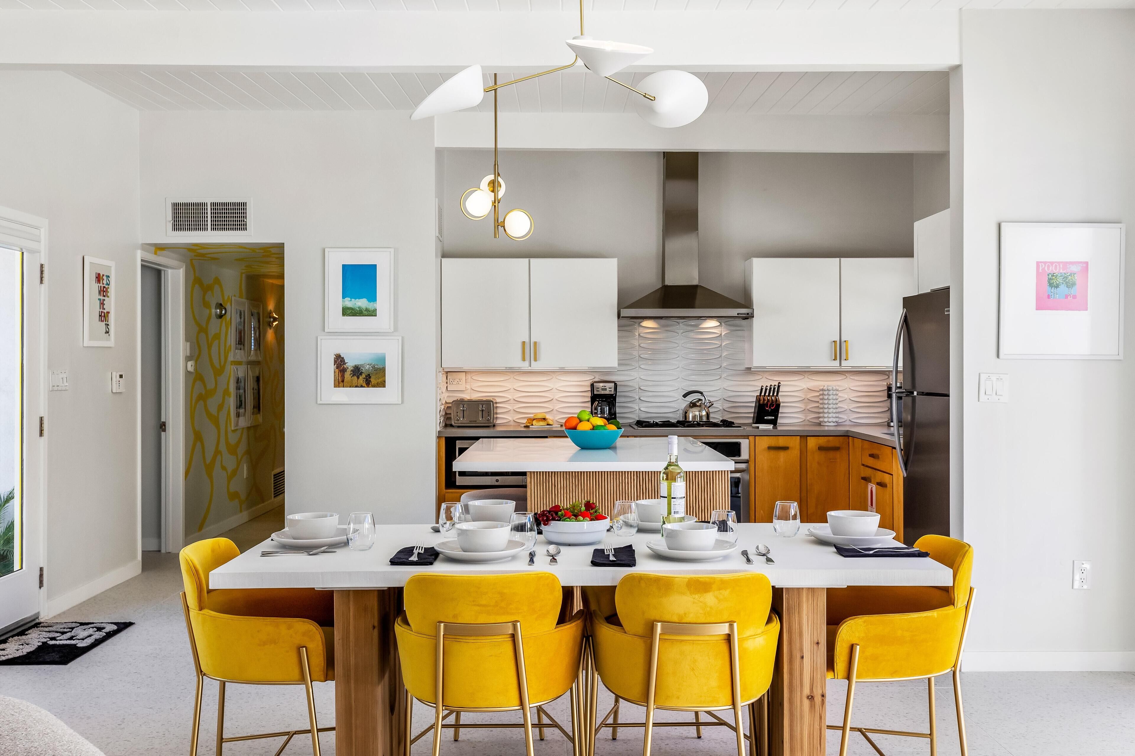 A modern kitchen with white cabinets, a central island with a bowl of fruit, and a dining area with yellow chairs and a white table.