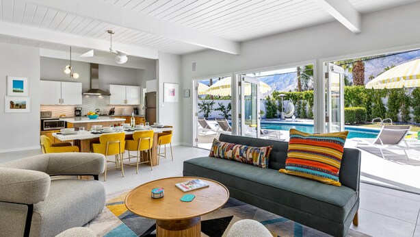 A modern living room with a sofa, armchairs, and a coffee table. There is a dining area with yellow chairs and a table. The room has large windows with a view of a pool and mountains. - Mid Mod Sunsation (Palm Springs)