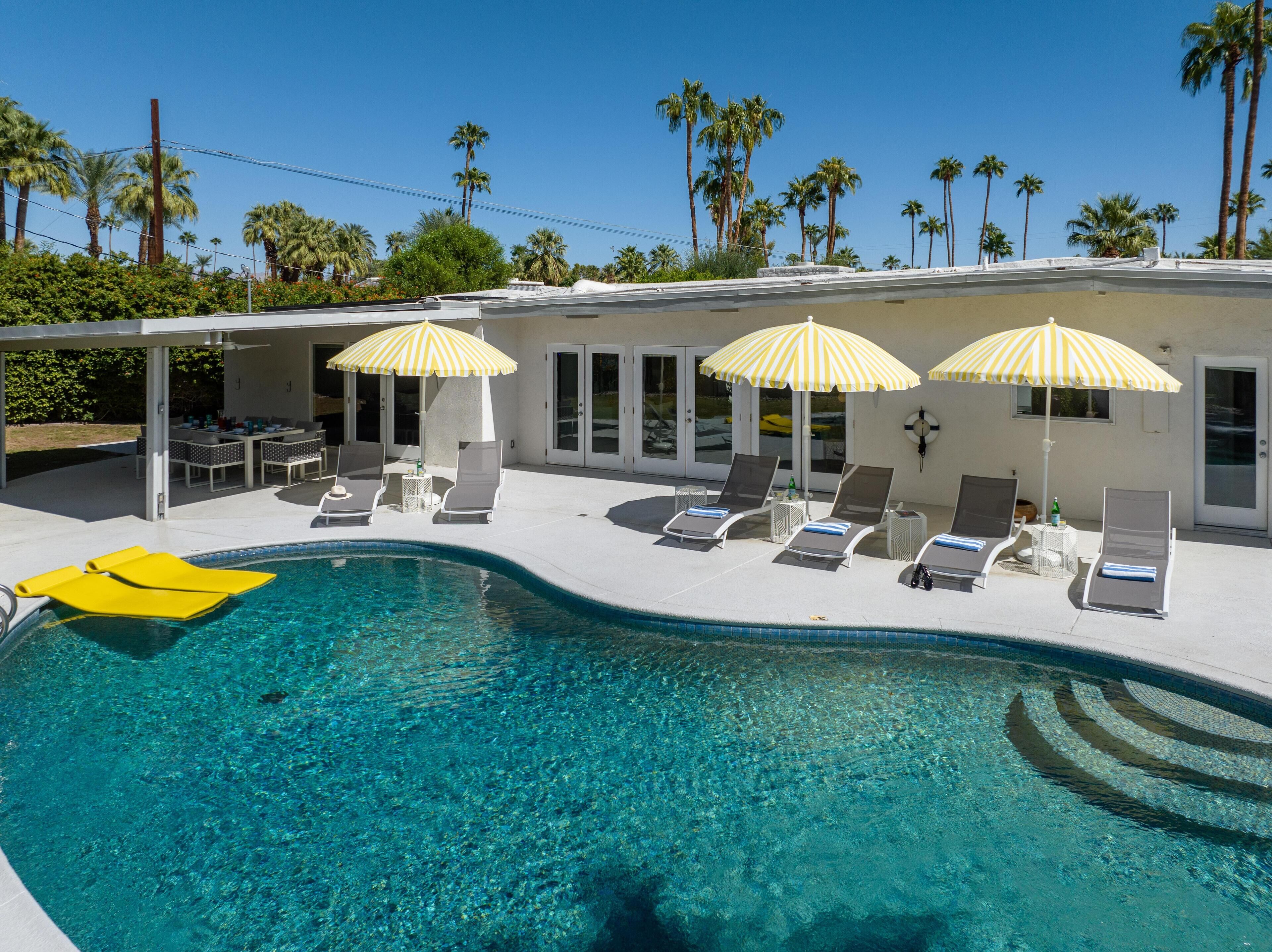 A poolside area with lounge chairs and umbrellas, a clear pool, and palm trees in the background.
