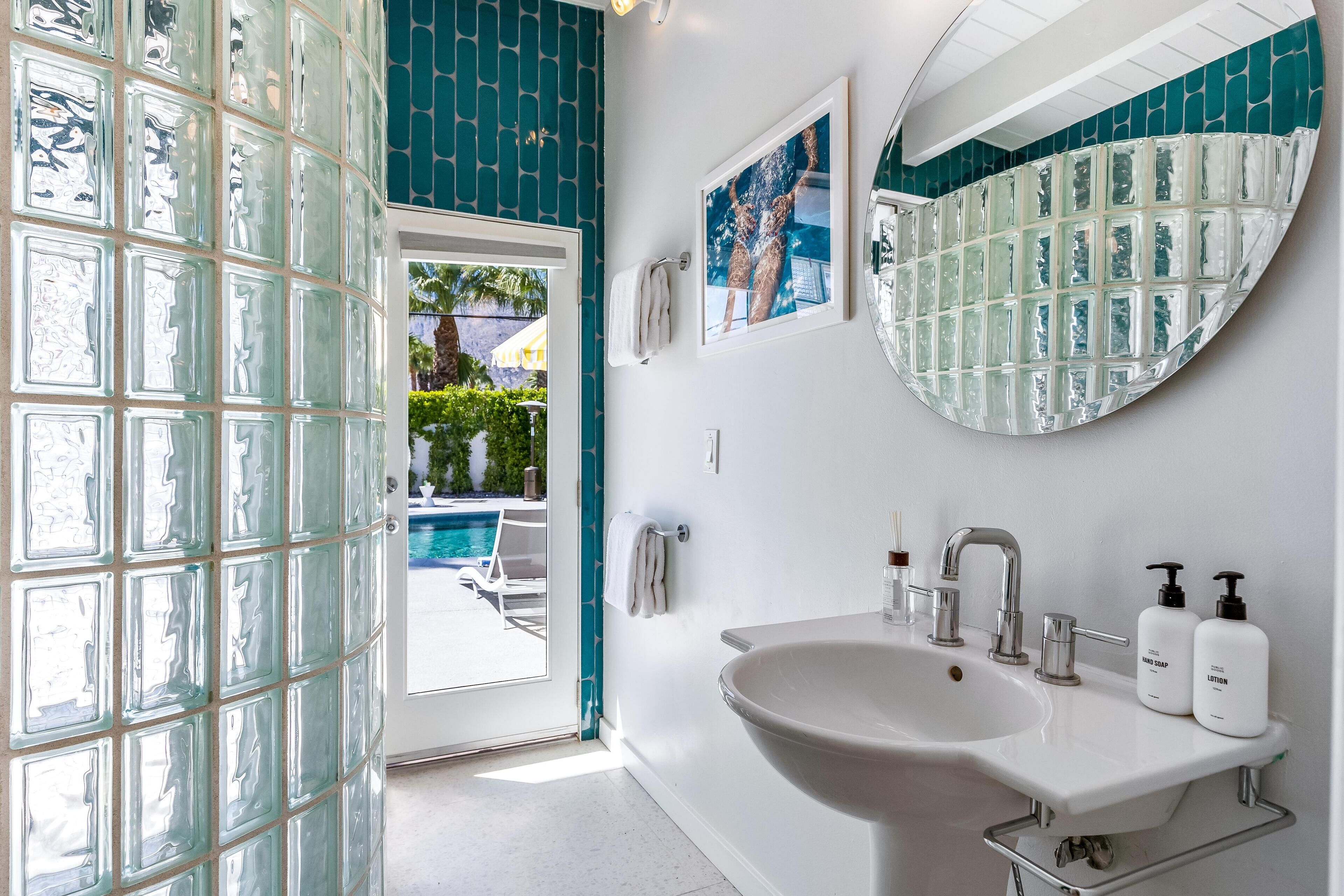 Un bany amb una gran porta de vidre, un lavabo blanc amb un mirall a sobre i dues ampolles de sabó de mans i loció al lavabo.