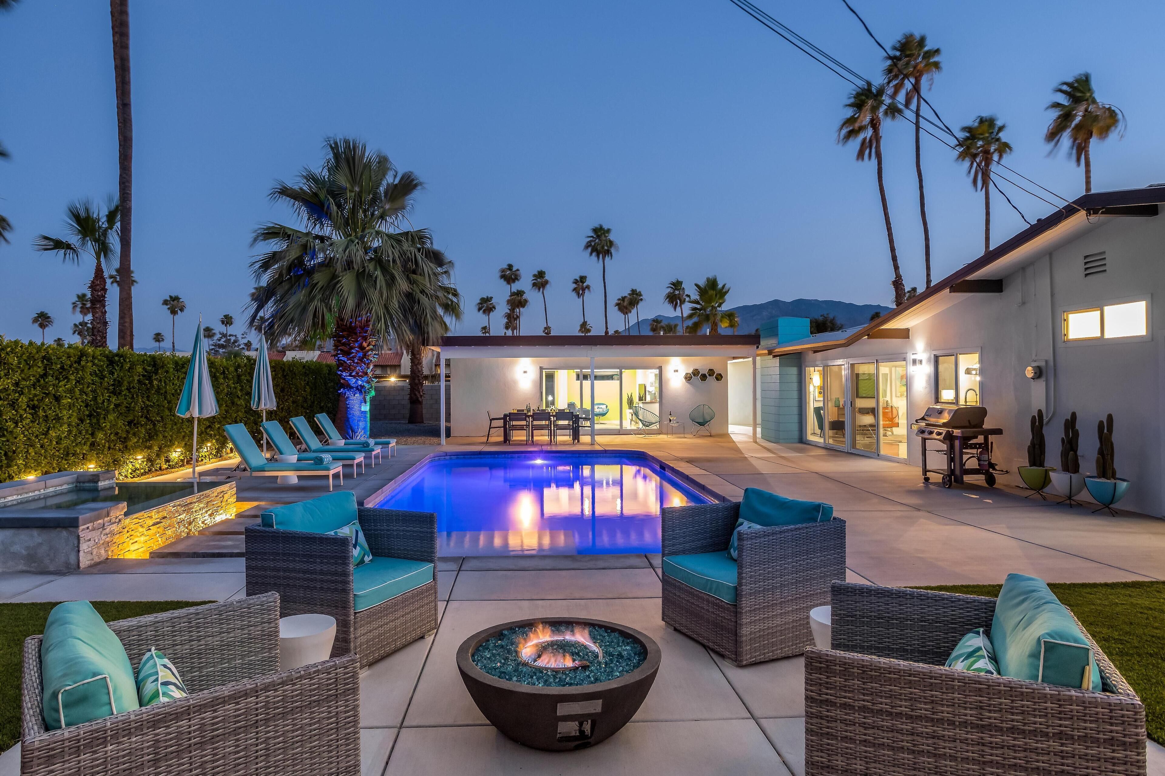 A pool area with lounge chairs and a fire pit, surrounded by palm trees and a modern house.