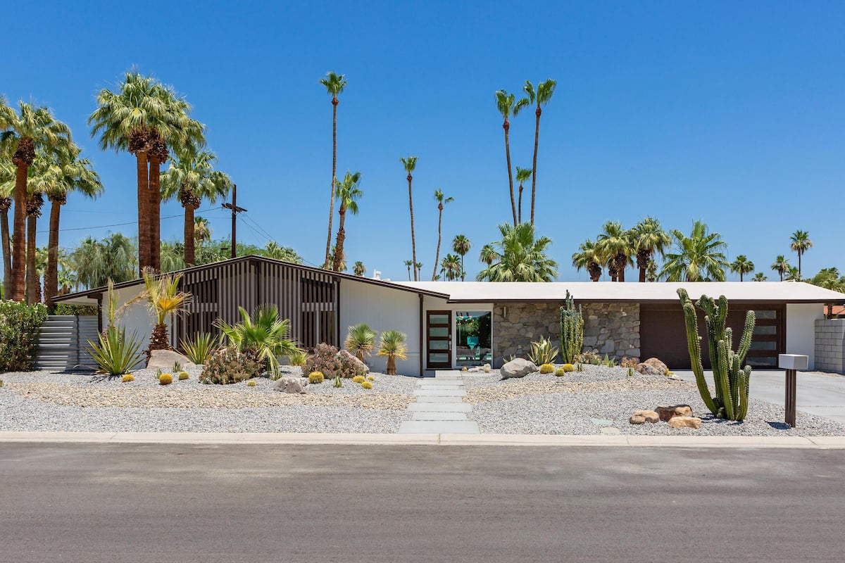 A modern house with a stone facade, surrounded by a desert landscape with tall palm trees and cacti.