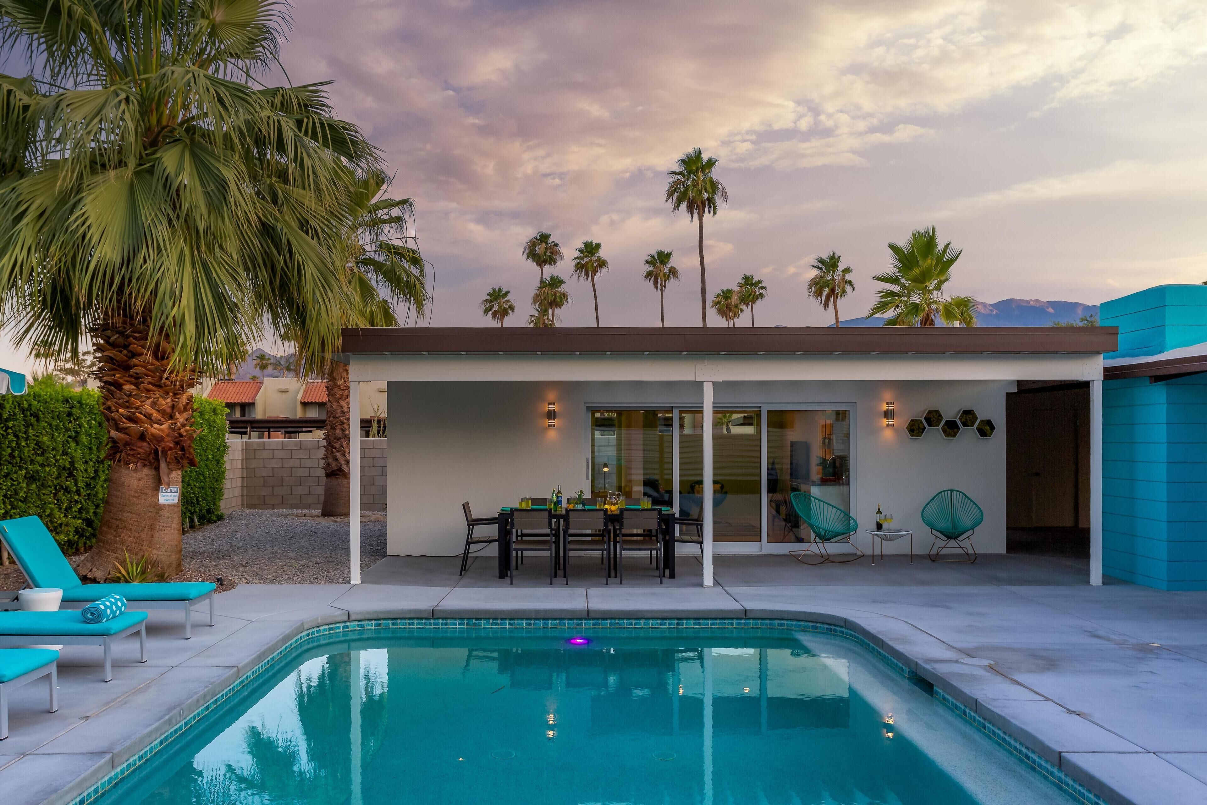 A modern outdoor pool area with a covered seating area, palm trees, and a clear view of the mountains in the background.