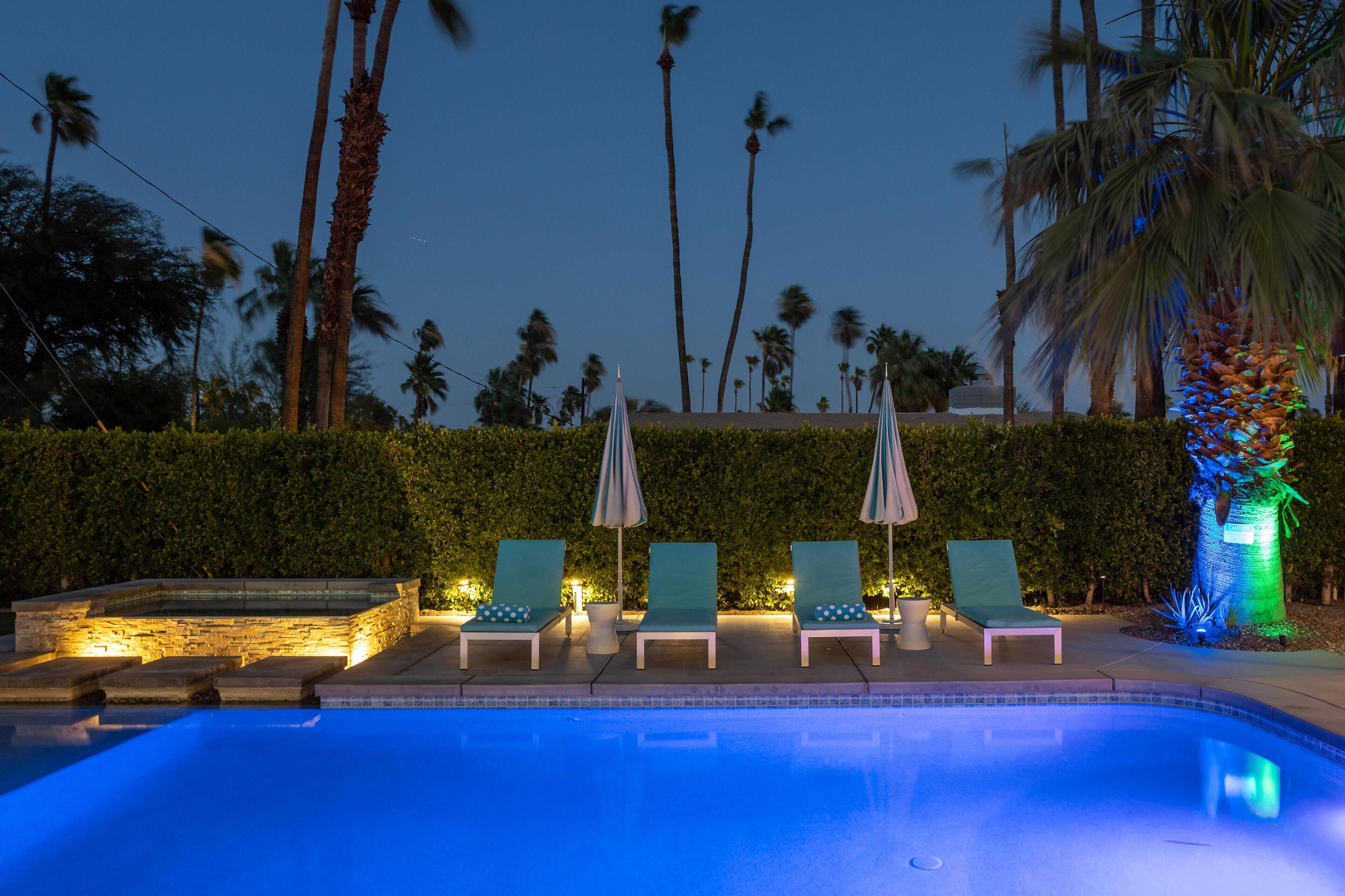A poolside area with lounge chairs and umbrellas, surrounded by palm trees and a hedge.