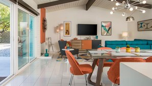 A modern living room with a large TV, teal sofa, orange chairs, and a glass dining table. - Alexander Sunrise (Palm Springs)