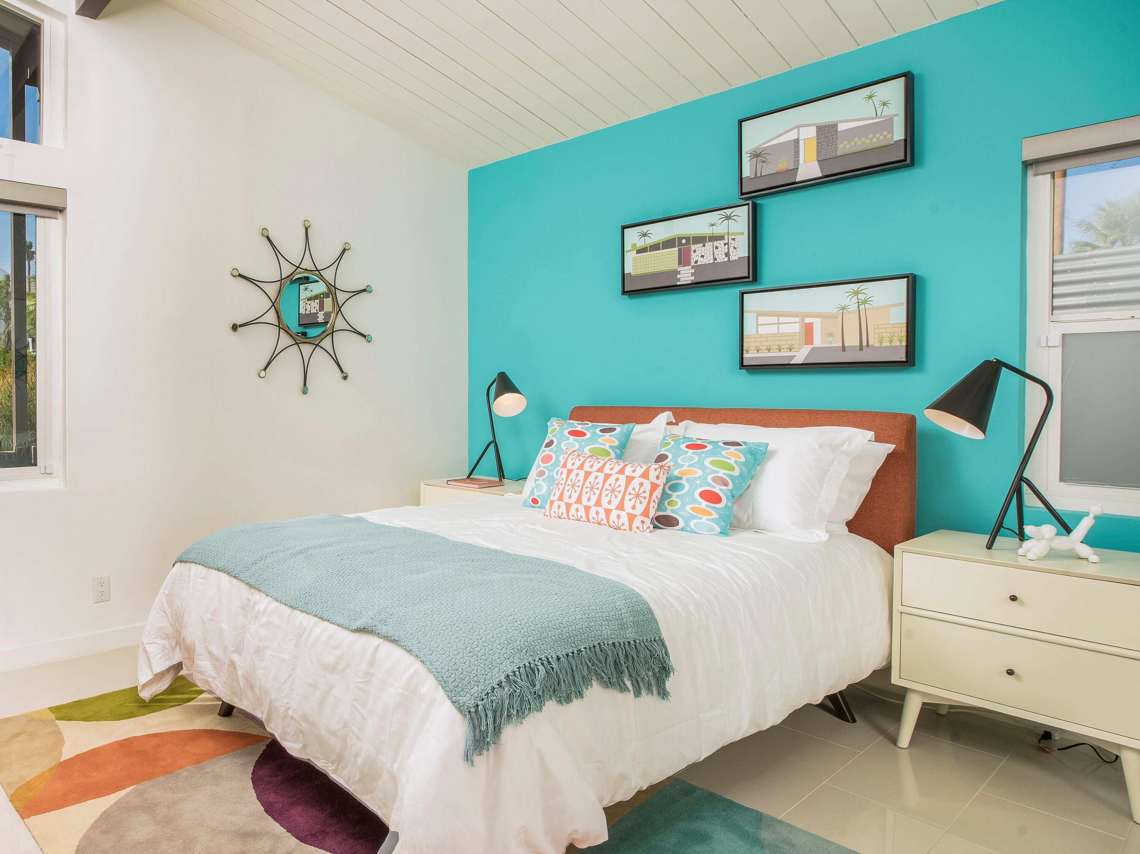 A bedroom with a teal accent wall, a bed with colorful pillows, a nightstand with a lamp, and framed pictures on the wall.