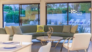 A modern living room with a green sofa, white chairs, and a glass coffee table. Large windows offer a view of a pool and outdoor furniture. - Mid-century Deluxe (Palm Springs)