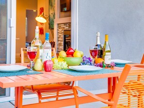 An outdoor dining area with a table set for two, featuring a variety of fruits, bottles of wine, and a decorative bowl.