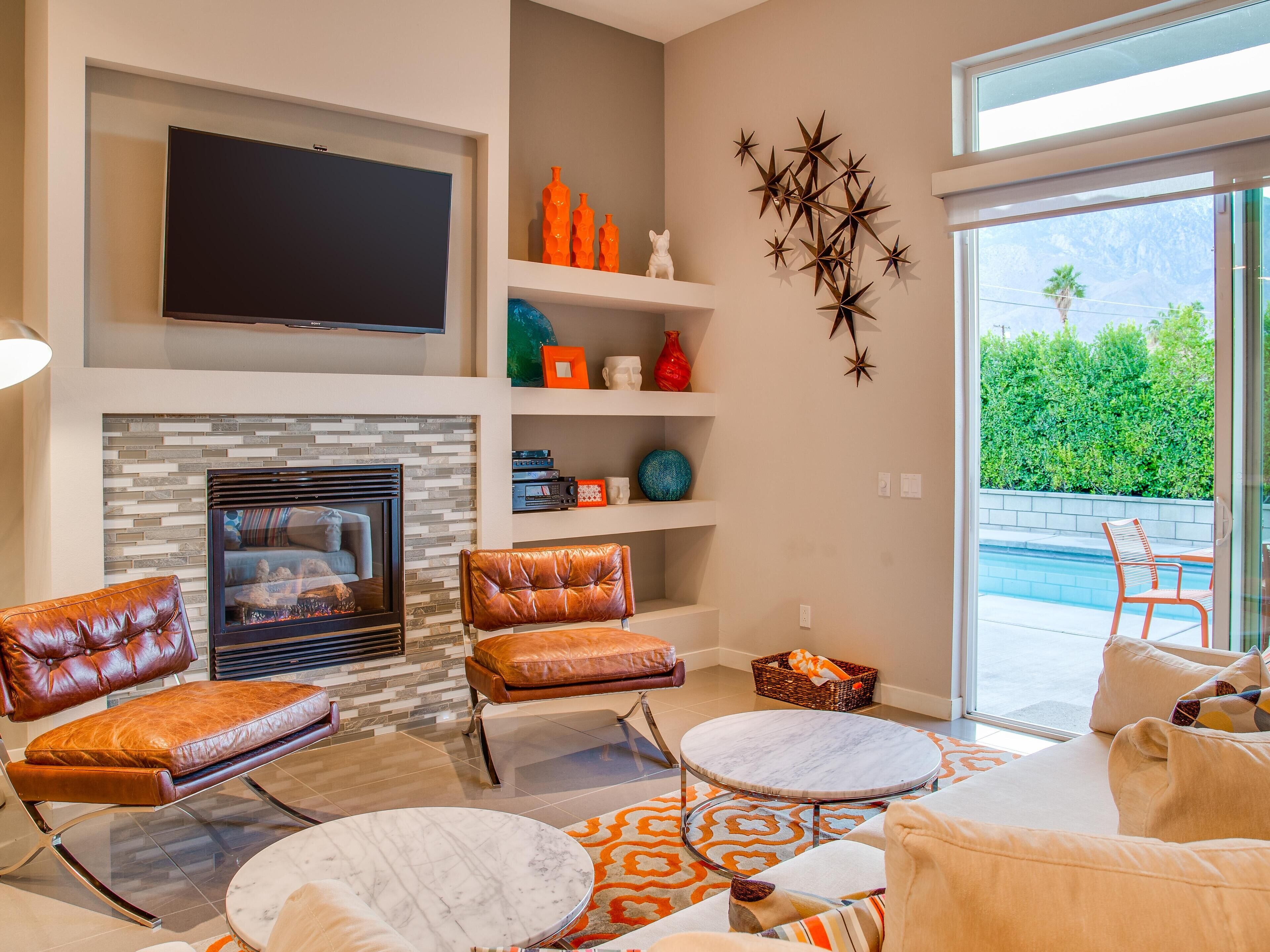 A modern living room with a fireplace, leather chairs, a TV, and a view of a pool.
