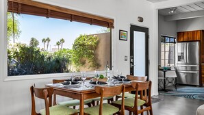 A dining area with a table set for four, wooden chairs with green cushions, a view of palm trees and greenery outside, and a modern kitchen with stainless steel appliances. - Lucy I'm Home (Palm Springs)