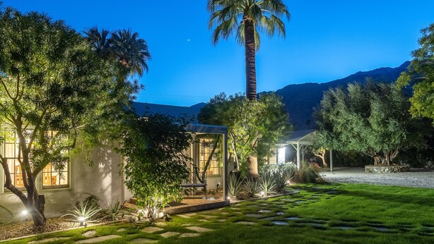 Um jardim bem iluminado com uma palmeira, um gazebo e uma casa com grandes janelas.